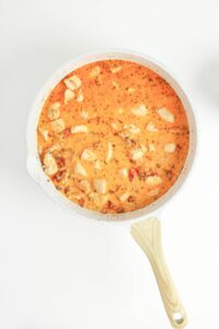 A white skillet filled with creamy orange sauce containing chunks of chicken and herbs, viewed from above on a white background.