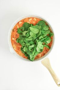 A white skillet with a creamy tomato-based sauce, chunks of chicken, and a layer of fresh spinach leaves on top.
