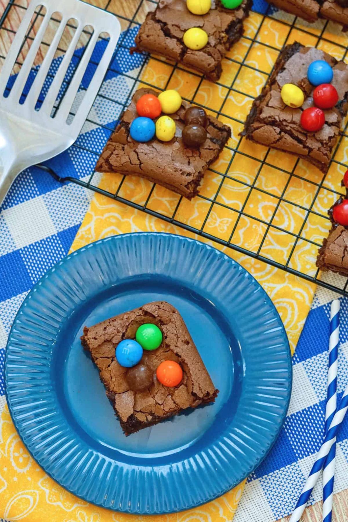 A brownie square topped with colorful candy-coated chocolates on a blue plate, with more brownies on a wire rack in the background.
