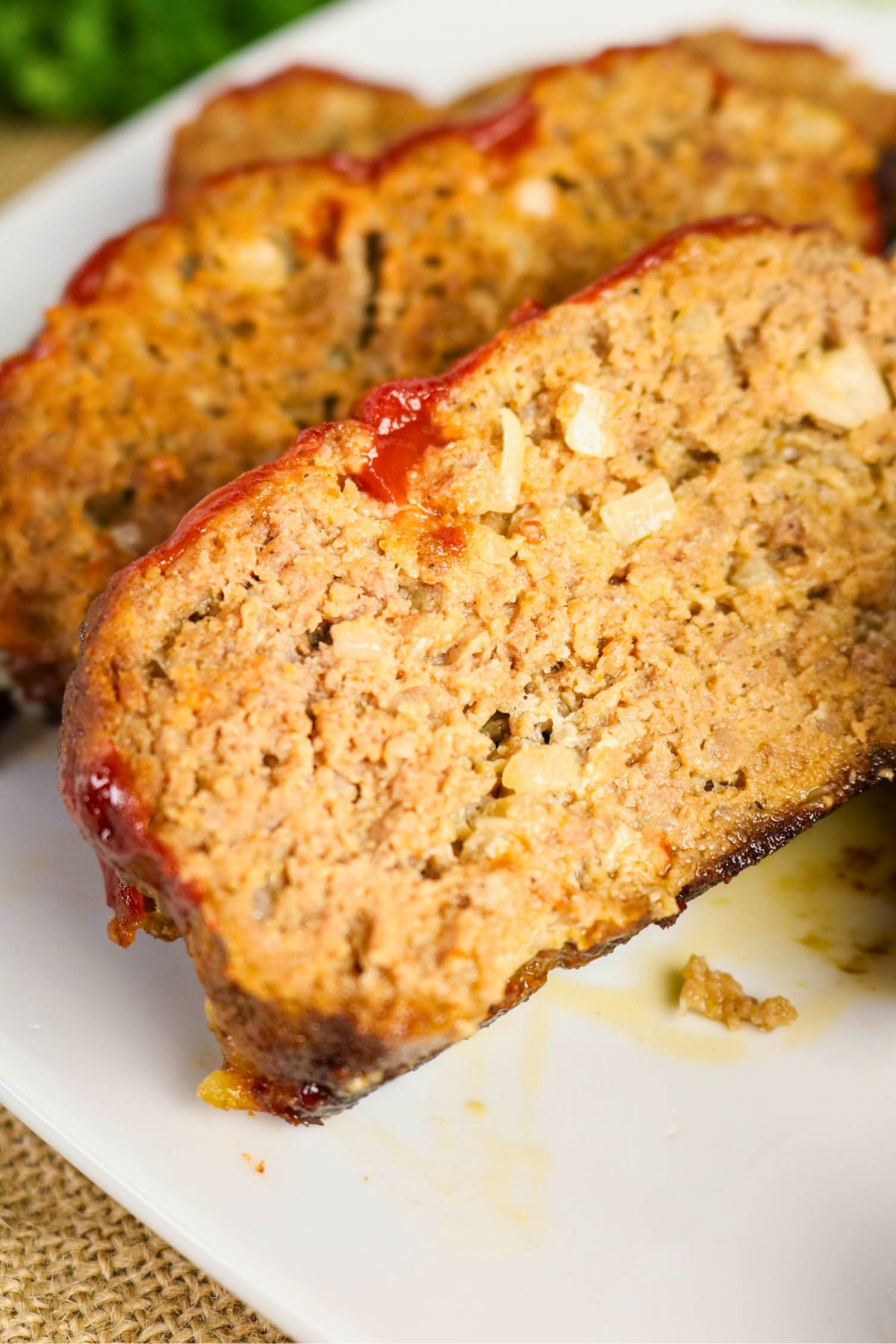 Close-up of sliced meatloaf on a white plate, showing a moist texture with visible bits of onion and a layer of ketchup glaze on top.