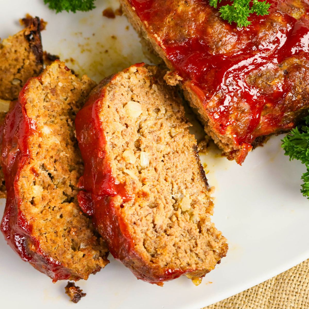 Two slices of meatloaf with a tomato glaze are served on a white plate, garnished with parsley.