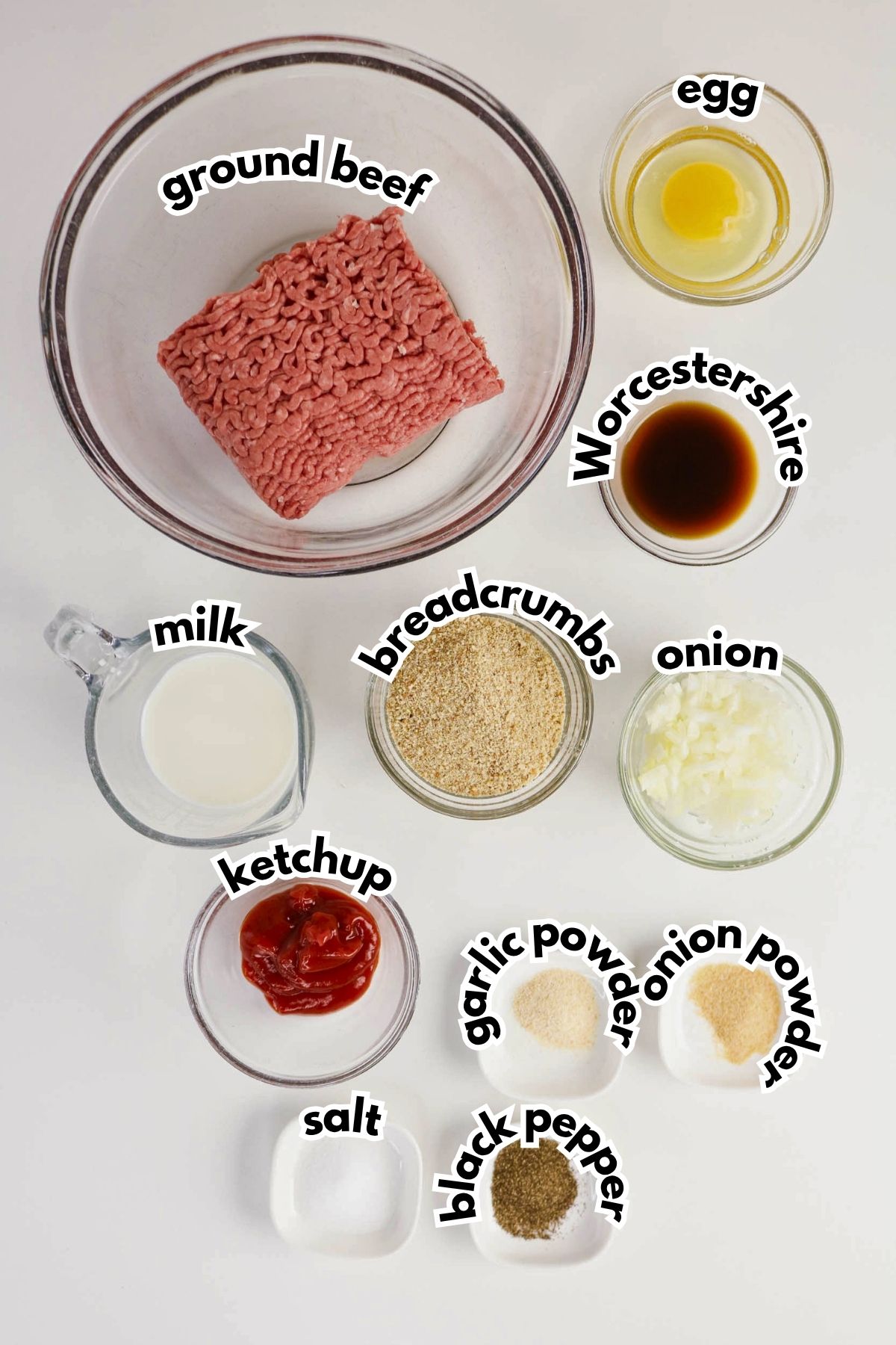A top view of bowls containing ground beef, egg, Worcestershire sauce, milk, breadcrumbs, onion, ketchup, garlic powder, onion powder, salt, and black pepper.