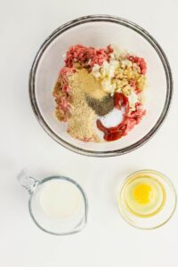 A glass bowl with ground meat, chopped onions, garlic, breadcrumbs, ketchup, and spices. Next to it are a small glass pitcher of milk and a bowl with a raw egg.