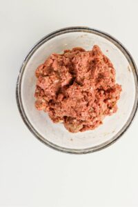 A clear glass bowl filled with raw ground meat mixture sits on a white surface.