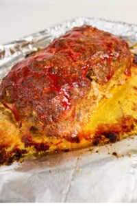 A baked meatloaf topped with tomato sauce sits on a foil-lined baking sheet, showing browned edges and some rendered juices.