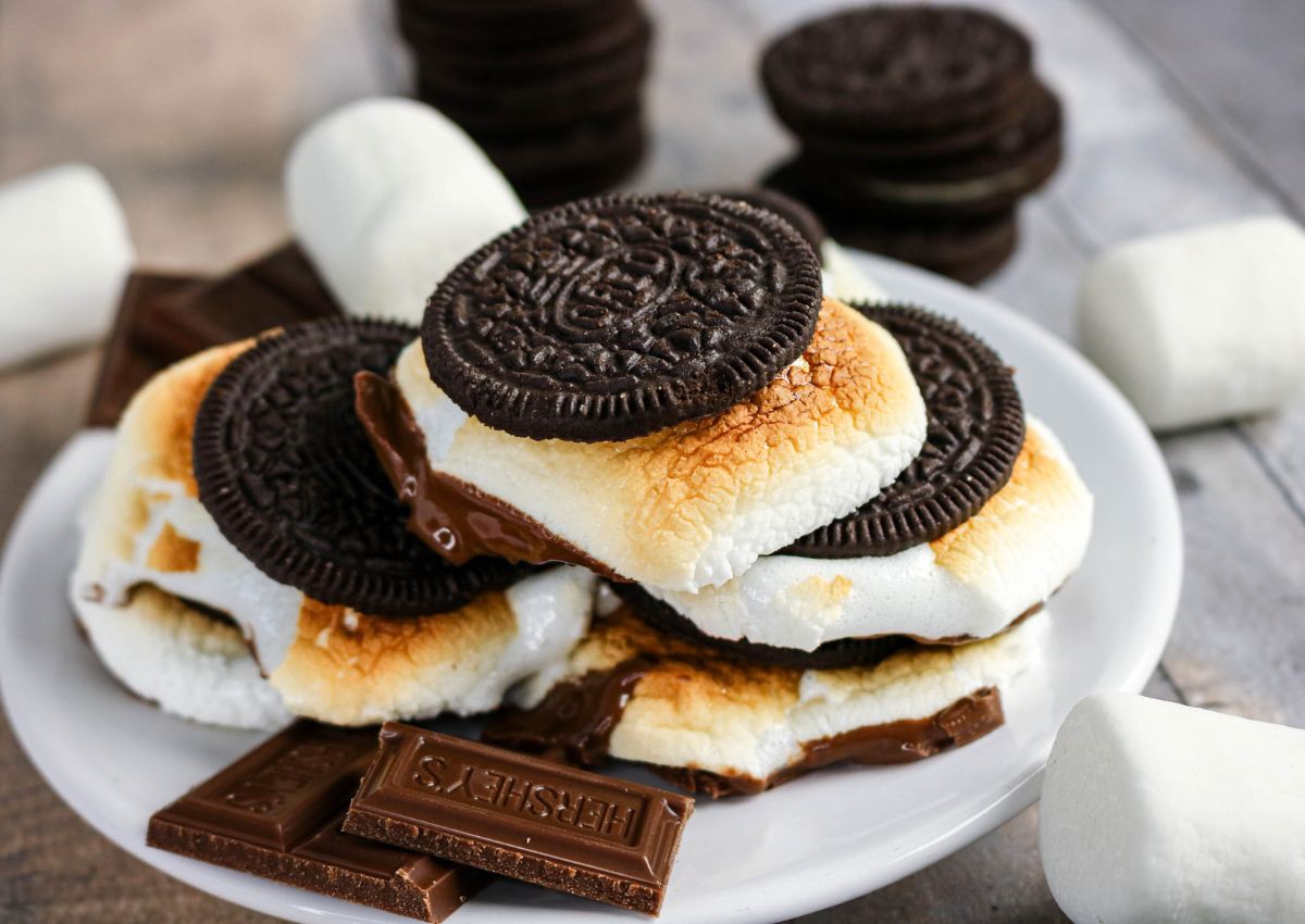 Plate of smores made with toasted marshmallows, chocolate, and chocolate sandwich cookies, with extra marshmallows and chocolate pieces nearby.