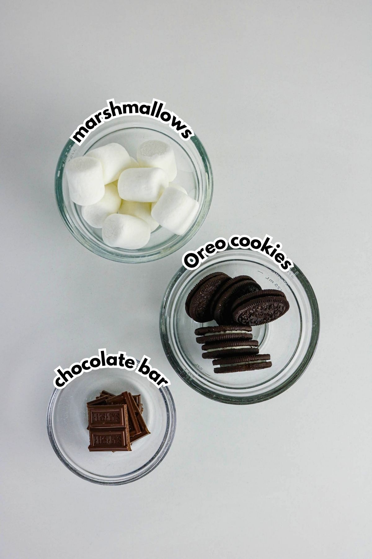 Three glass bowls containing marshmallows, Oreo cookies, and pieces of chocolate bar are labeled on a white surface.