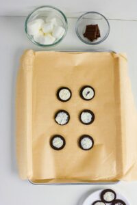 A baking sheet lined with parchment paper holds eight chocolate sandwich cookies with cream exposed. Glass bowls contain marshmallows and chocolate squares nearby.