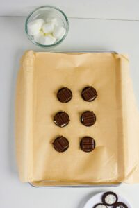 A baking sheet lined with parchment paper holds six cookie sandwiches topped with chocolate squares; a bowl of marshmallows and a plate with cookies are nearby.