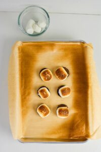 Six toasted smores sit on a parchment-lined baking sheet, with a glass bowl containing three marshmallows above them on a white surface.