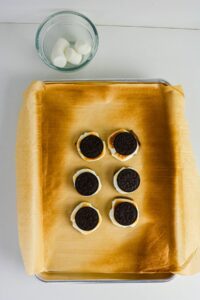 Six Oreo and marshmallow treats on a parchment-lined baking tray, with a glass bowl containing marshmallows in the background.