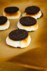 Chocolate sandwich cookies placed on toasted marshmallows, arranged on a sheet of brown parchment paper.