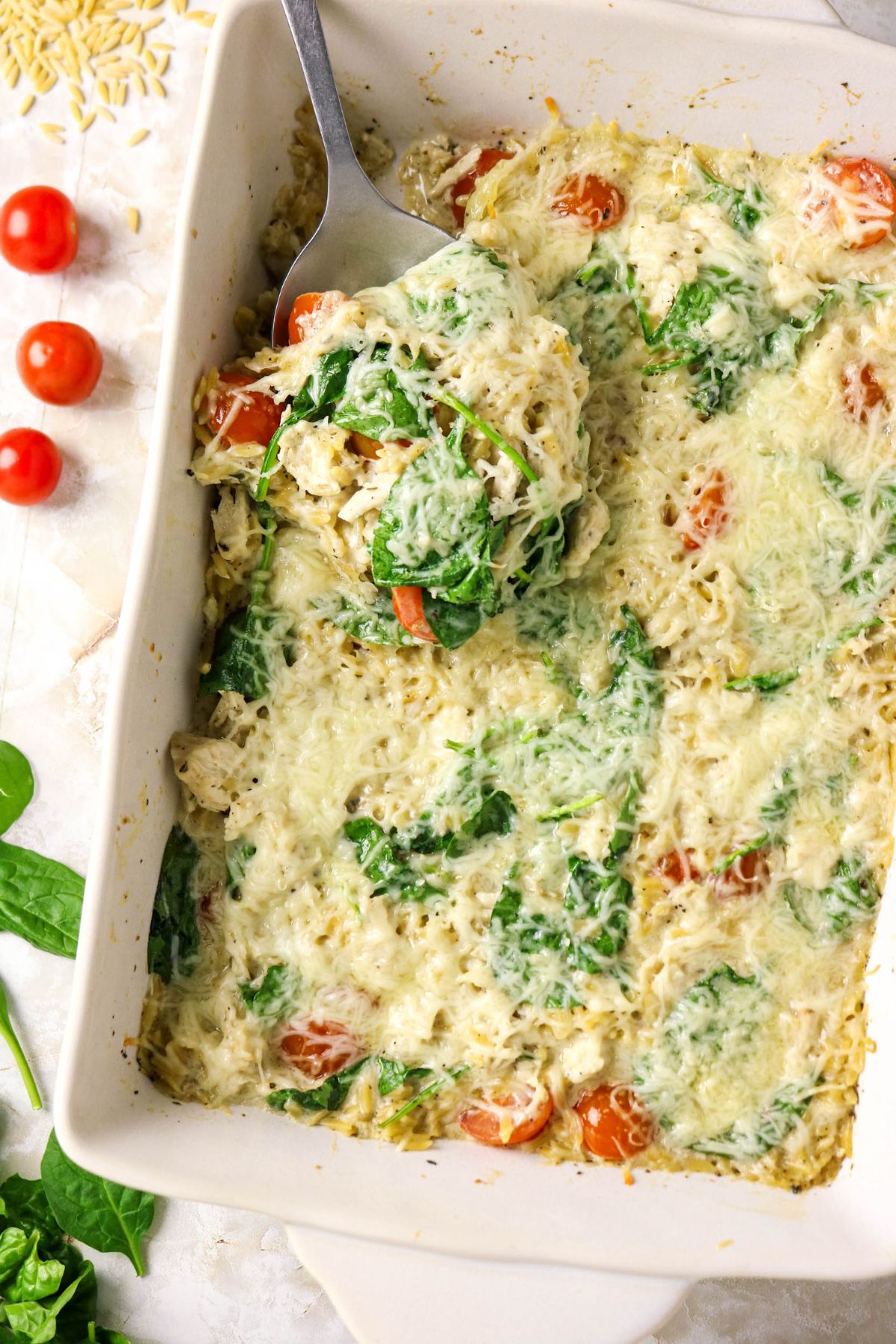 A baked casserole in a white dish with melted cheese, spinach, and cherry tomatoes, with a serving spoon lifting a portion.