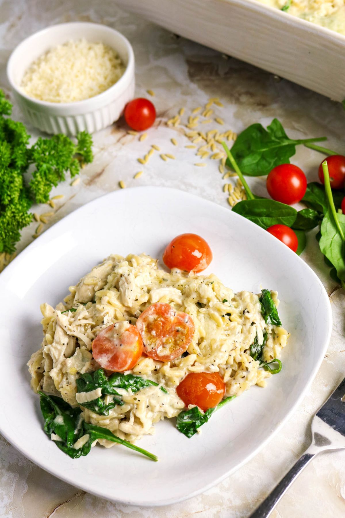 A plate of creamy pasta with spinach and cherry tomatoes, garnished with herbs, sits on a marble surface; grated cheese and vegetables are visible in the background.