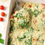 A spoon scoops a portion of a baked spinach, tomato, and cheese casserole from a white dish, with fresh spinach leaves and cherry tomatoes nearby.