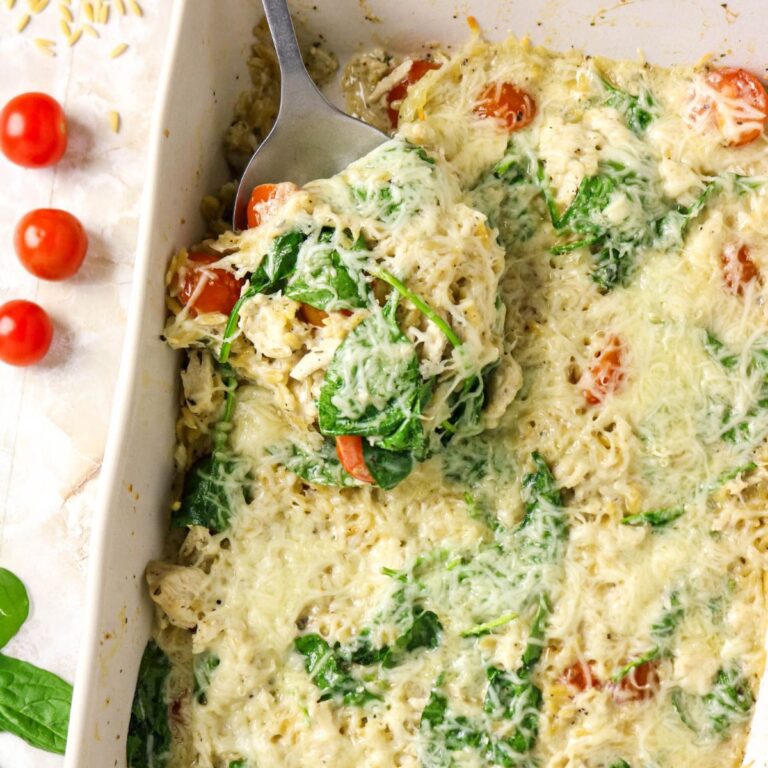 A spoon scoops a portion of a baked spinach, tomato, and cheese casserole from a white dish, with fresh spinach leaves and cherry tomatoes nearby.