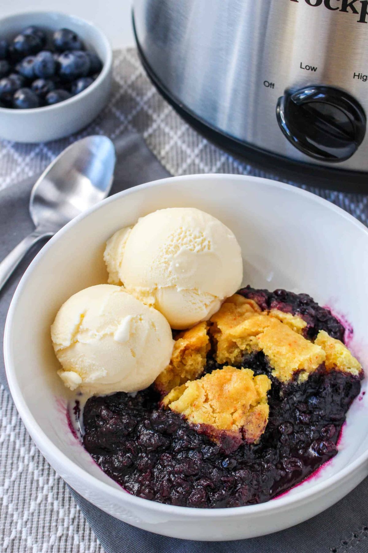 A bowl of blueberry cobbler topped with two scoops of vanilla ice cream, placed next to a spoon and a slow cooker.