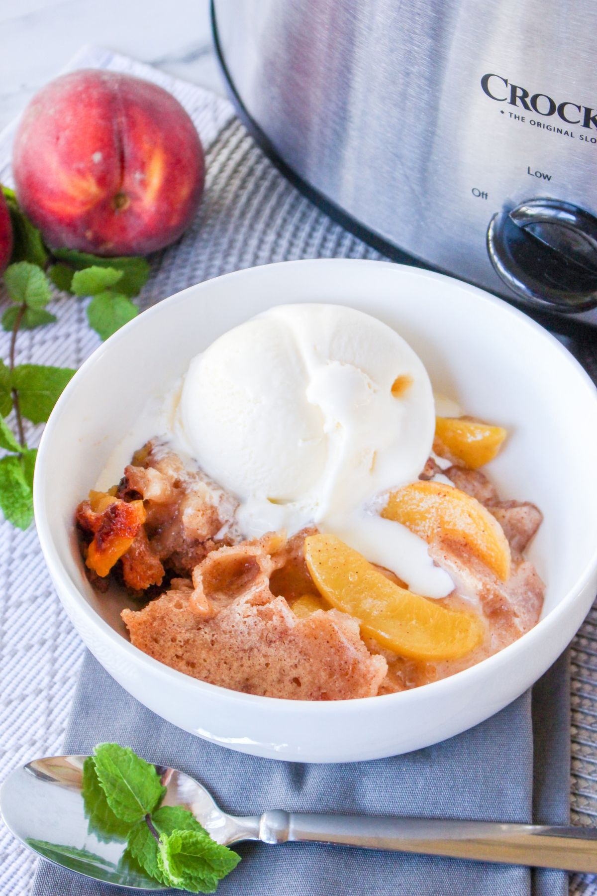 A bowl of peach cobbler topped with a scoop of vanilla ice cream, placed next to a slow cooker, a peach, and a spoon on a napkin.