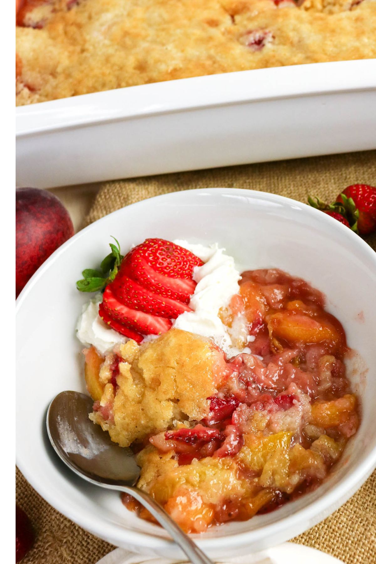 A bowl of peach and strawberry cobbler topped with whipped cream and sliced strawberries, with a spoon resting inside; a baking dish is in the background.