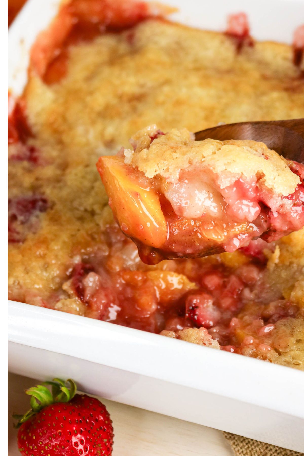 A close-up of a baked fruit cobbler being served with a wooden spoon from a white dish, with visible peaches and strawberries.