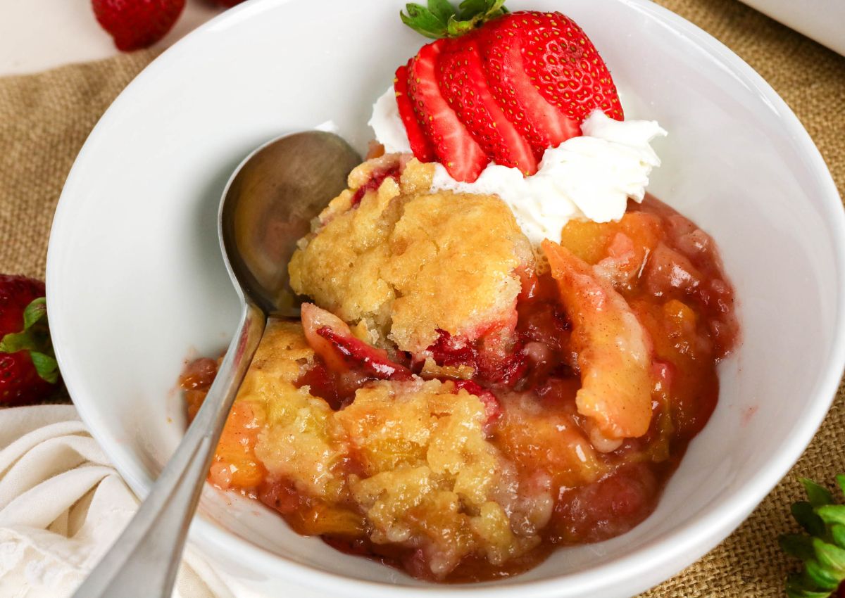 A bowl of peach and strawberry cobbler topped with whipped cream and sliced strawberries, with a spoon resting inside.