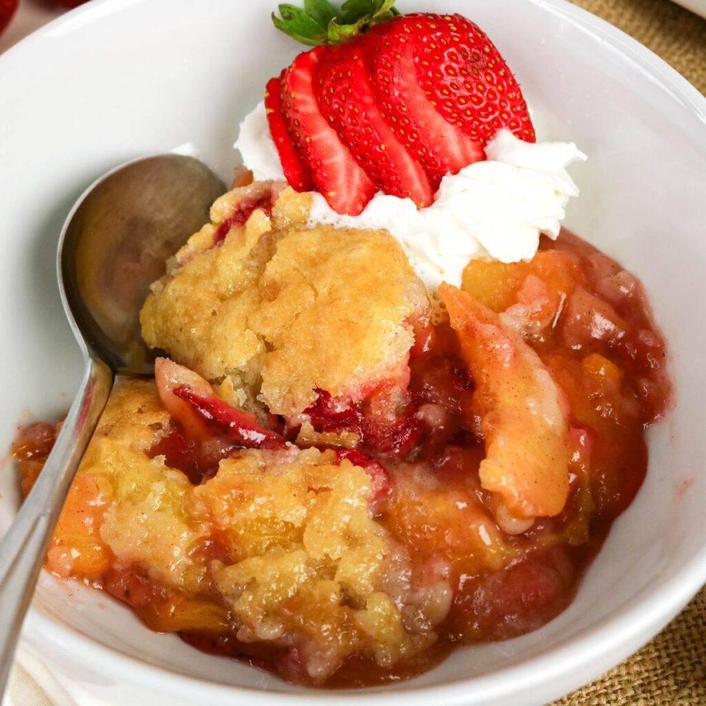 A bowl of fruit cobbler topped with whipped cream and sliced strawberries, with a spoon resting inside the bowl.