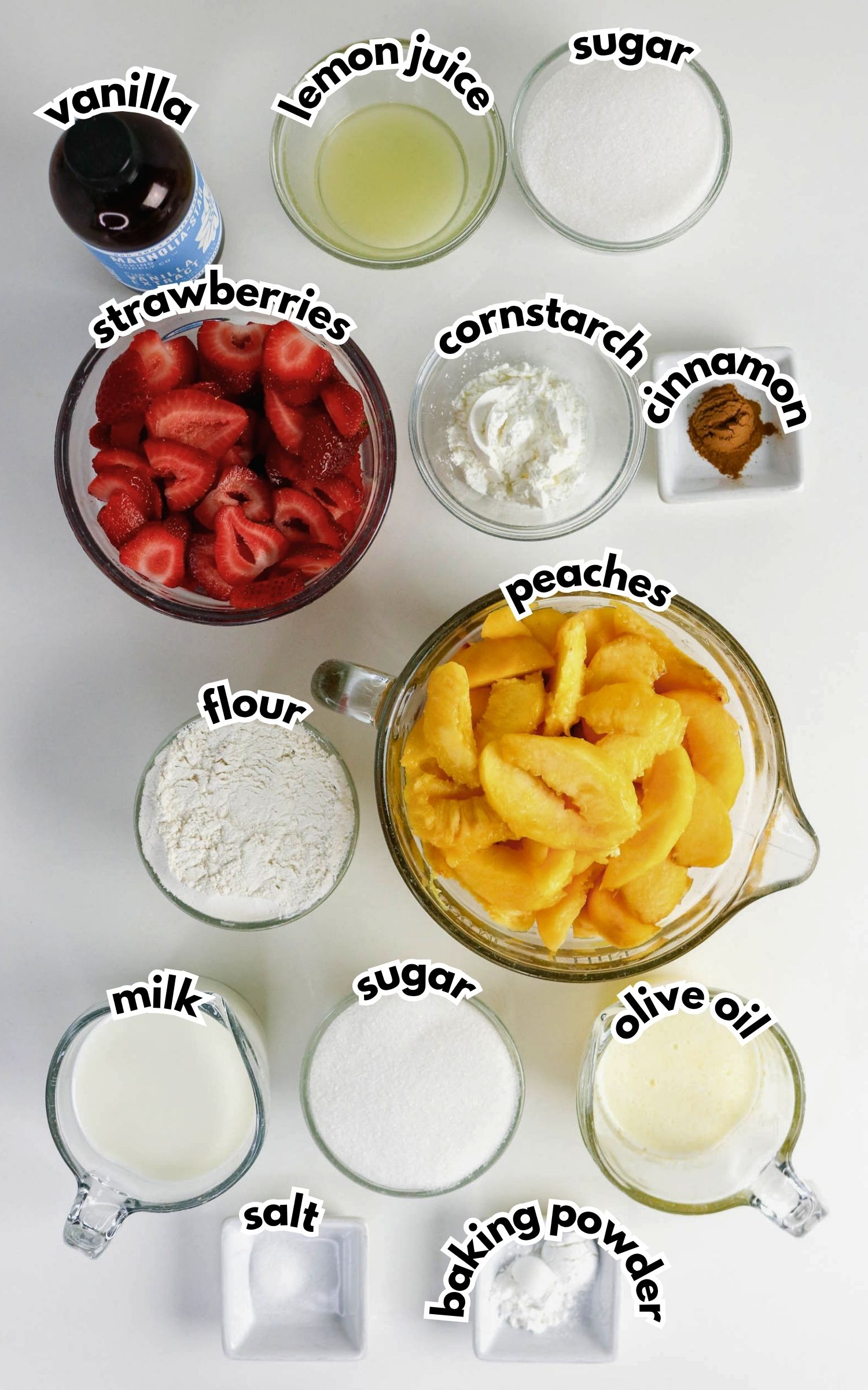 Overhead view of bowls and measuring cups containing strawberries, peaches, flour, sugar, cornstarch, cinnamon, vanilla, milk, olive oil, salt, lemon juice, and baking powder.