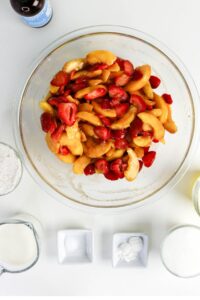 A glass bowl with sliced peaches and strawberries is surrounded by bowls and cups of flour, milk, sugar, oil, and baking ingredients on a white surface.