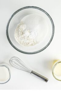 A glass bowl with flour, sugar, and baking powder, a metal whisk, a glass of milk, and a bowl of melted butter on a white surface.