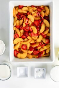 A baking dish filled with sliced peaches and strawberries, surrounded by small bowls of flour, milk, sugar, and baking ingredients on a white surface.