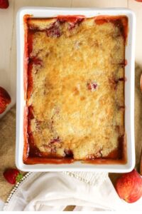 A baked strawberry cobbler in a rectangular white dish, with a golden-brown crust and fruit filling visible around the edges.