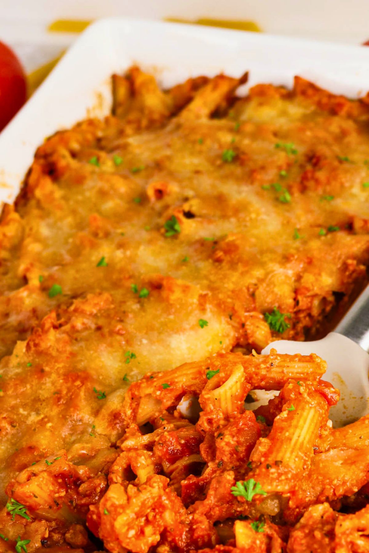 A close-up of a baked pasta casserole topped with melted cheese, with a serving spoon lifting out a portion.