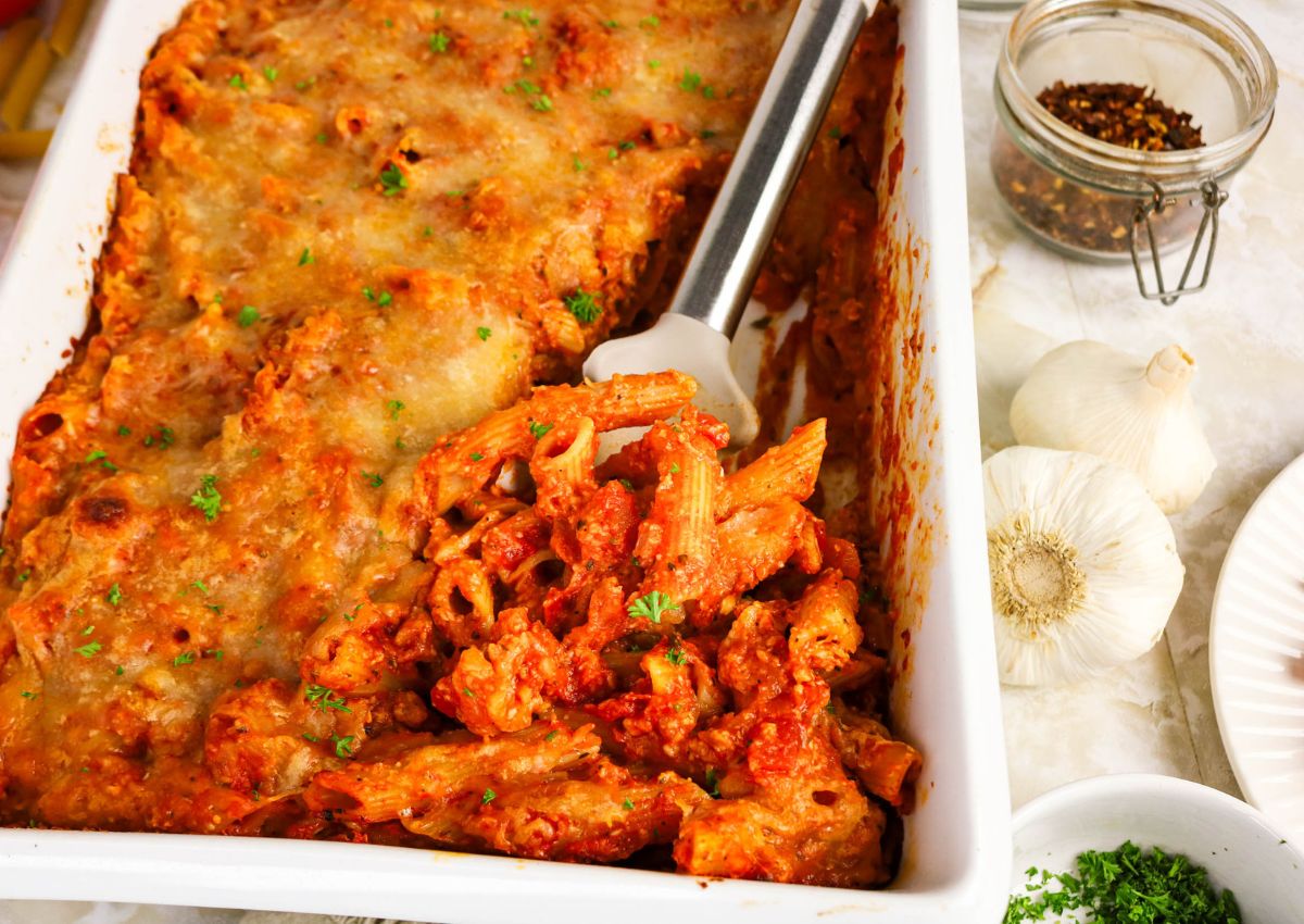 A baked pasta dish with melted cheese and tomato sauce in a white casserole dish, served with a spatula. Garlic bulbs, red pepper flakes, and herbs are nearby.