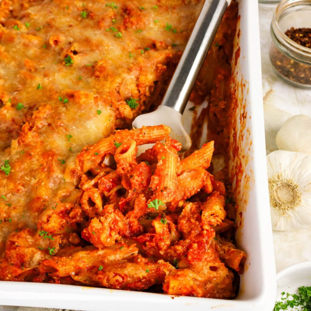 Baked pasta casserole with melted cheese on top, partially served with a spatula in a white dish; garlic bulbs and red pepper flakes are visible on the side.