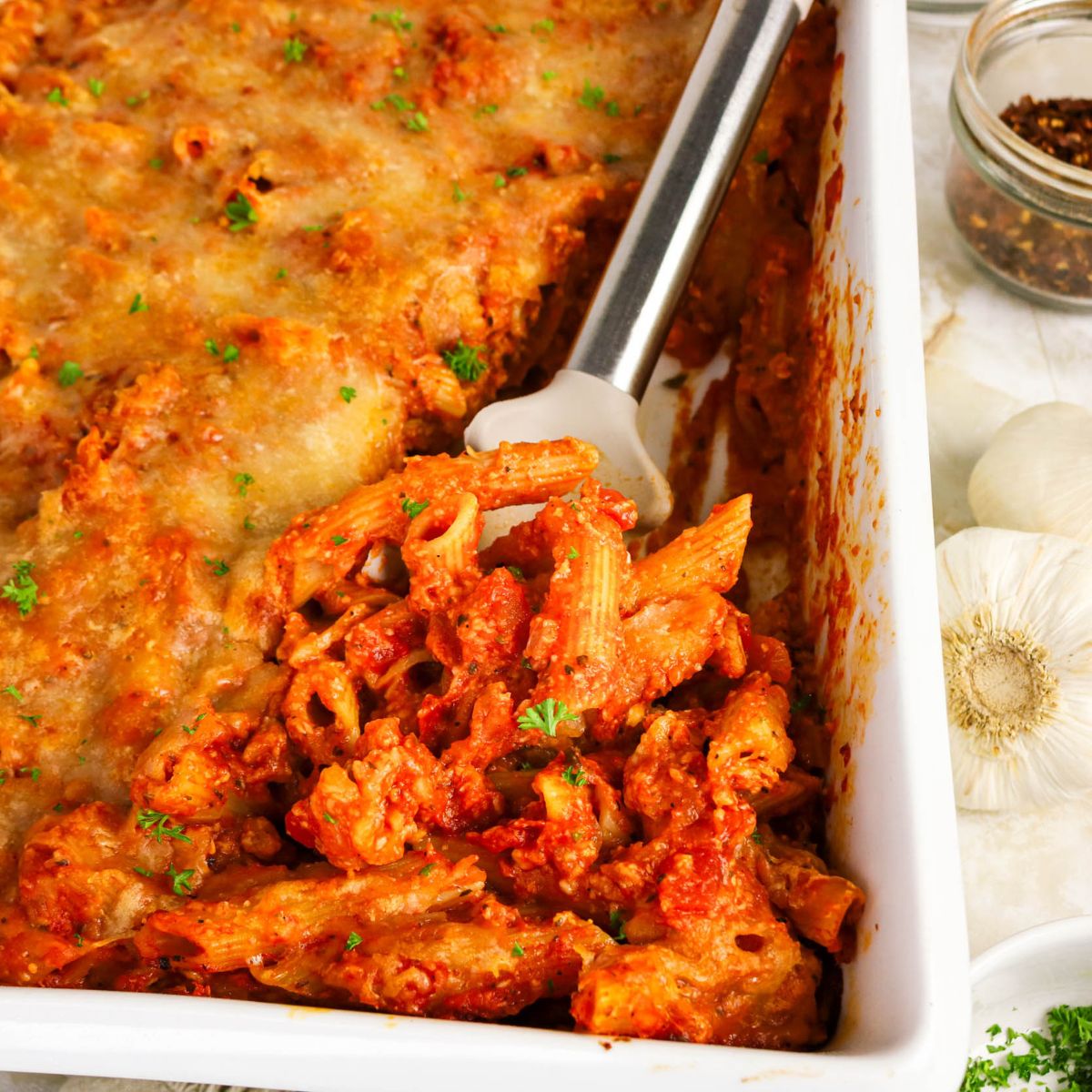Baked pasta casserole with melted cheese on top, partially served with a spatula in a white dish; garlic bulbs and red pepper flakes are visible on the side.