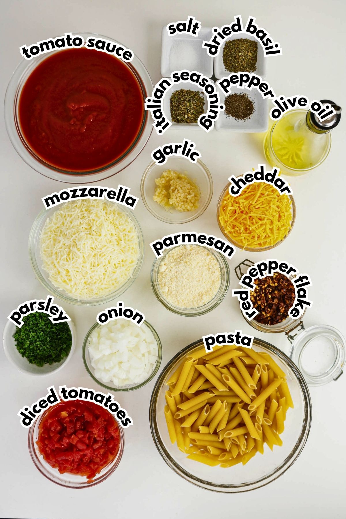 Various ingredients for pasta bake are arranged in bowls, including pasta, cheeses, tomato sauce, diced tomatoes, spices, herbs, garlic, onion, olive oil, and red pepper flakes.