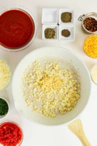 A white skillet with chopped onions and garlic sits on a countertop surrounded by bowls of tomato sauce, cheese, herbs, diced tomatoes, and spices.