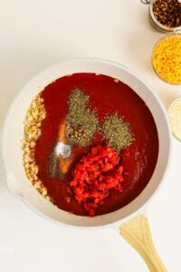 A skillet with tomato sauce, diced tomatoes, chopped onions, and assorted spices is shown on a white surface next to small bowls of cheese and other ingredients.