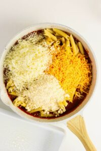 A white skillet filled with uncooked penne pasta, tomato sauce, and three types of shredded cheese, viewed from above on a white surface.