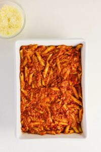 Rectangular white dish filled with penne pasta mixed with tomato meat sauce. A small bowl of shredded cheese is placed beside the dish.