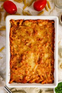 Baked pasta casserole topped with melted cheese in a rectangular white dish, surrounded by raw pasta, tomatoes, and herbs on a light countertop.