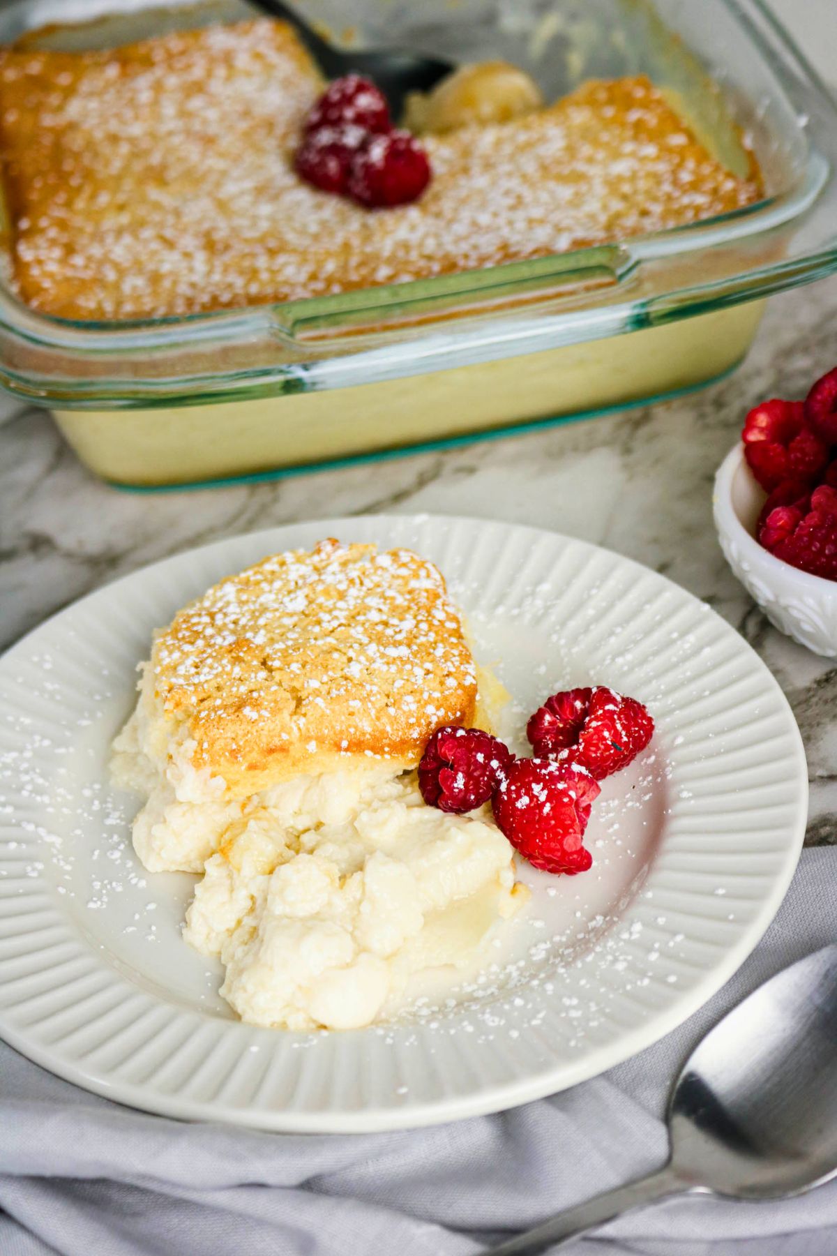 A piece of lemon pudding cake topped with powdered sugar is served on a white plate with fresh raspberries, next to a glass baking dish and a small bowl of raspberries.