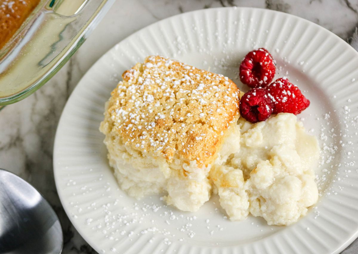 A serving of baked dessert topped with powdered sugar sits on a white plate, accompanied by three raspberries.