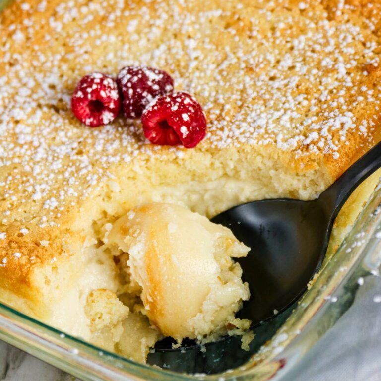 A glass baking dish with a serving of lemon pudding cake scooped out, topped with powdered sugar and three raspberries. A black spoon rests in the dish.