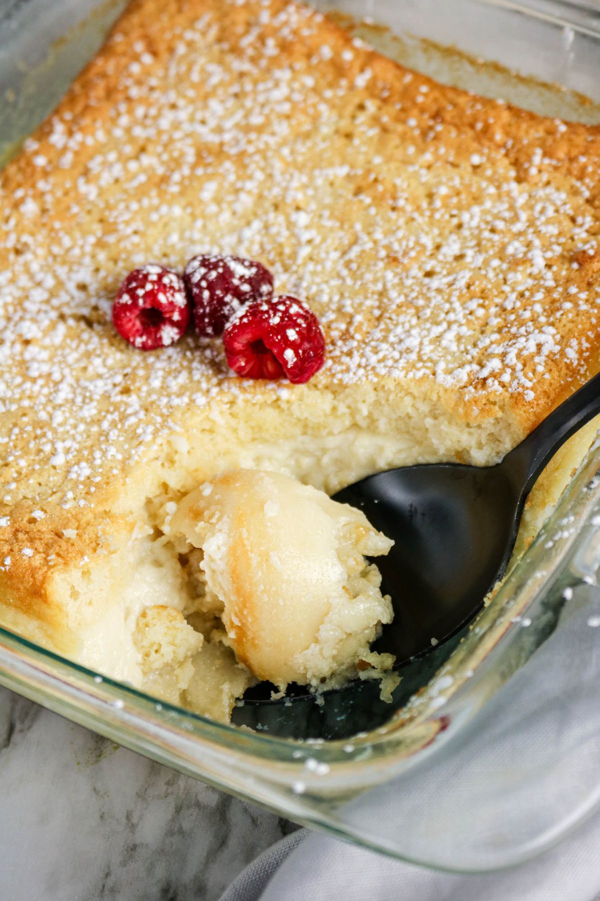 A glass baking dish with a partially served lemon pudding cake, topped with powdered sugar and three raspberries, with a black serving spoon inside.