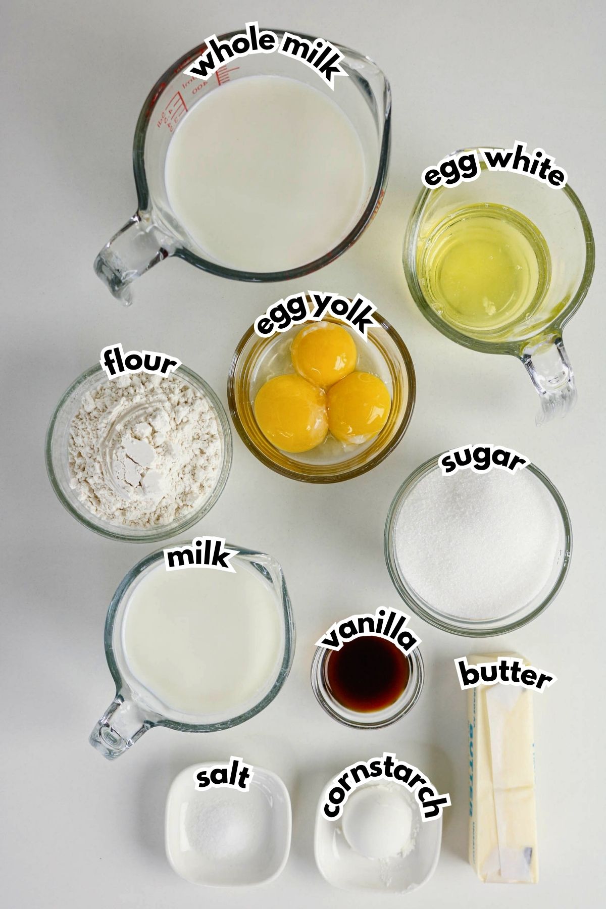 Top-down view of labeled ingredients in bowls and cups: whole milk, egg white, egg yolks, flour, milk, sugar, vanilla, butter, salt, and cornstarch on a white surface.