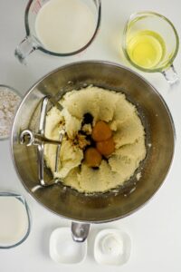 A mixing bowl with creamed butter and sugar, eggs, and vanilla extract, surrounded by bowls of flour, milk, egg whites, salt, and baking powder on a white surface.