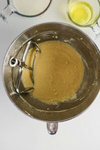 A mixing bowl with batter and a flat beater attachment, shown next to a measuring cup of milk and a measuring cup of oil on a white surface.
