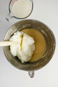 A metal mixing bowl with yellow batter and whipped egg whites being folded in, next to a measuring cup of milk on a white surface.