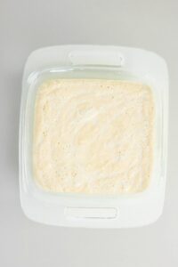 A glass baking dish filled with a frothy, pale yellow liquid mixture, viewed from above on a light gray surface.
